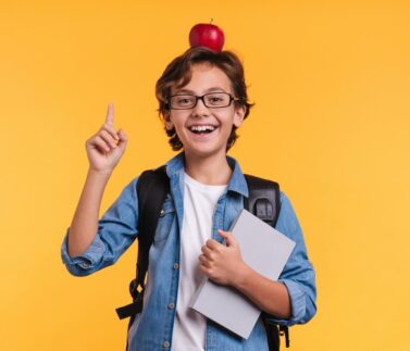 Smart male kid pointing at copy space holding books for school and apple on his head isolated over yellow background