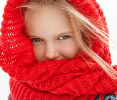 Close-up portrait of beautiful European blonde female school child wearing red warm wrap scarf covering her lips having walk outdoors in city park in sunny morning, enjoying winter vacations